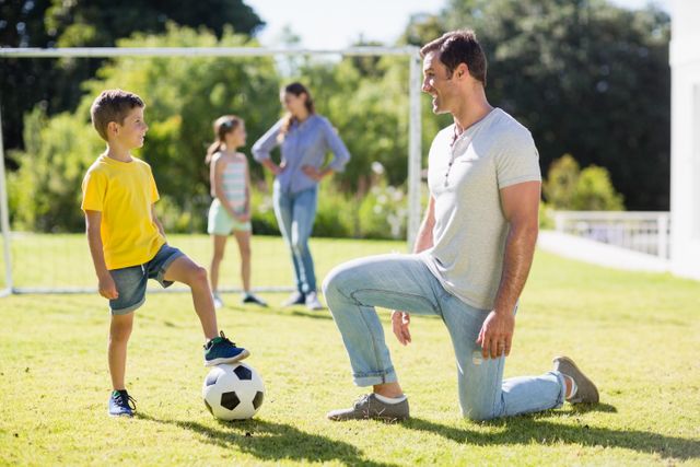 Father and son are enjoying a playful football game in the park on a beautiful sunny day. In the background, a woman and another child are standing and watching. This image is perfect for advertisements or articles related to family bonding, outdoor activities, sports, parenting, and promoting a healthy lifestyle. It exemplifies the joy and connection experienced during family activities.