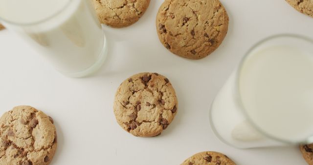 Flat lay featuring freshly baked chocolate chip cookies arranged alongside glasses of milk on white background. This image is ideal for food blogs, recipe websites, bakery advertisements, and dessert menus. Use it to evoke feelings of comfort and indulgence.