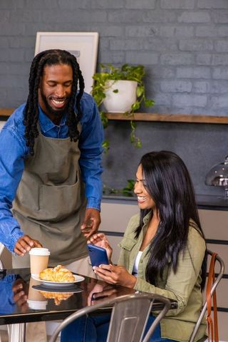 Smiling african american barista handing coffee to indian woman using phone at cafe
