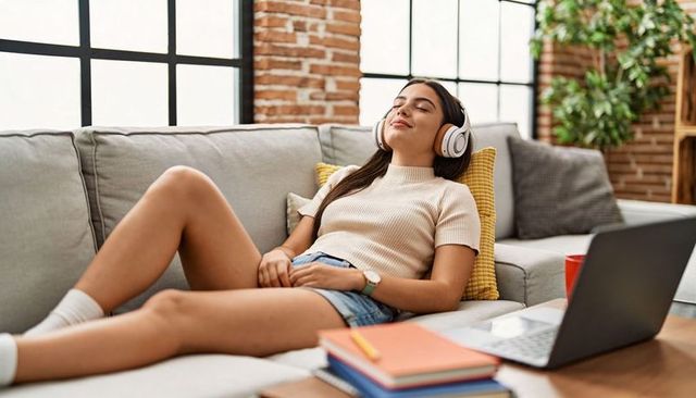 Relaxing young woman listening to music with headphones on sofa, laptop and notebooks nearby