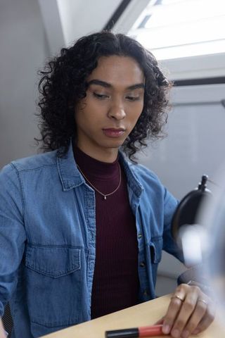 Curly-haired person holding lipstick at sunlit loft desk, denim shirt, contemplative mood
