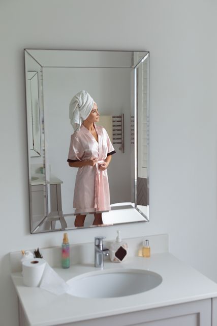 This image shows a woman tying the knot of her nightwear while looking at her reflection in the bathroom mirror. She has a towel wrapped around her head, suggesting she has just taken a shower. This photo can be used for articles or advertisements related to personal care, morning routines, self-care, and home lifestyle. It is ideal for promoting bath products, skincare routines, or home decor.