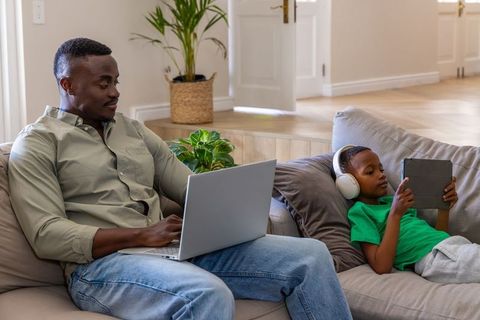 African american father working from home while son using tablet and headphones on sofa