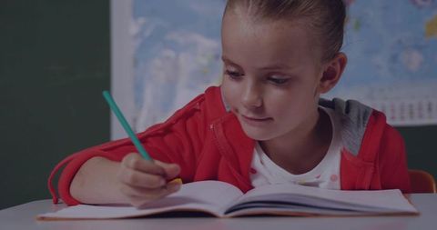 Young girl writing and concentrating in red hoodie at school desk with green pencil