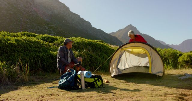 Senior man and woman enjoy camping adventure, setting up tent in beautiful mountain location with lush greenery on a sunny day. Great for depicting retirement lifestyle, outdoor recreation, natural scenery, travel, and adventurous activities. Useful for illustrating themes of health, wellness, and connection with nature.