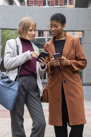 Professional women comparing smartphones in urban plaza wearing coats and business attire