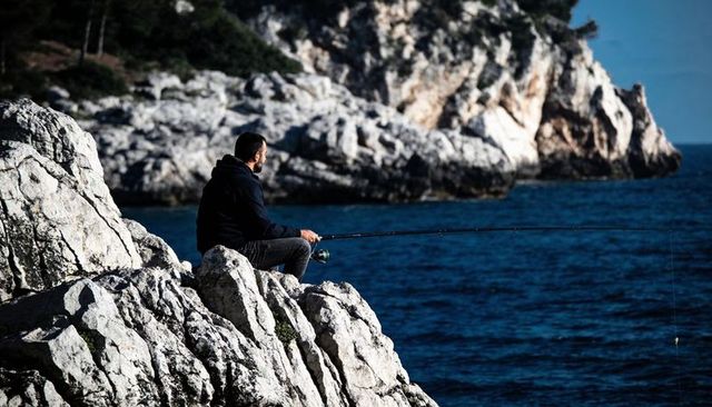Solitary angler sitting on limestone cliff fishing with spinning rod over deep blue sea