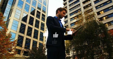 Businessman tapping tablet with floating thumbs-up among urban skyscrapers and trees