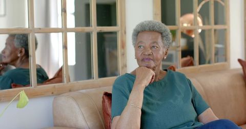 Senior african american woman sitting on tan sofa gazing into mirror, contemplative home