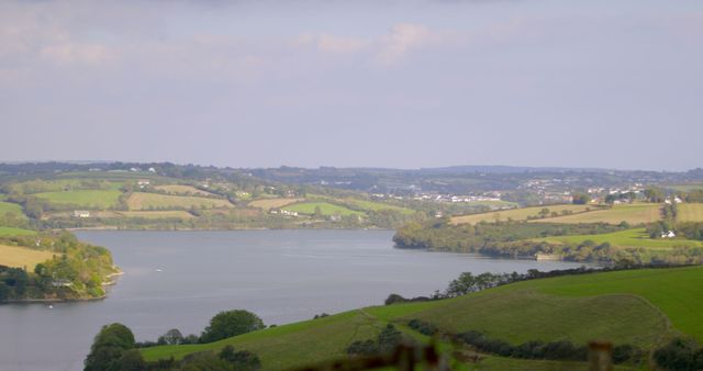Depicts tranquil river bend flanked by lush rolling hills and expansive farmland, with distant countryside homes. Ideal for nature-loving websites, blogs about rural life, travel guides promoting countryside tourism, or backgrounds in print and digital publications.