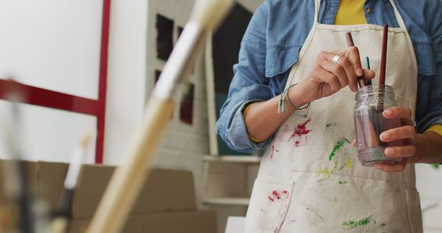 Image of midsection of biracial female artist in apron holding jar with brushes in studio. Art, crafts, creativity and creation process concept.