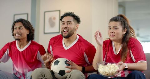 Friends cheering and holding soccer ball and popcorn while watching match on couch
