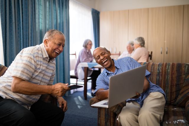 Happy senior friends looking at laptop while sitting on sofa against females in nursing home