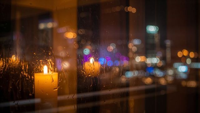 Flickering pillar candle reflecting on rain-splashed window with warm city bokeh glow