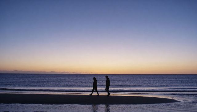 Two adults walking at dusk along tidal beach with silhouetted reflections and calm horizon