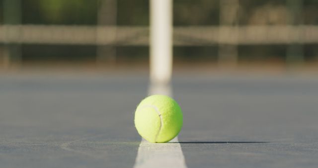 Tennis ball positioned precisely on court line with net subtly blurred in background. Ideal for use in sports promotions, training programs, recreational activity ads and tennis-focused content.
