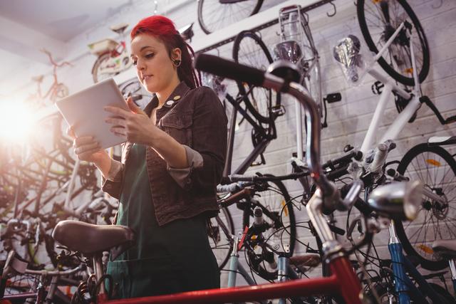 Young female mechanic in bicycle workshop using a digital tablet. She is surrounded by various bicycles and tools, focusing on her work. Ideal for use in industry news, marketing materials for bike shops, or articles about modern technology in traditional professions.