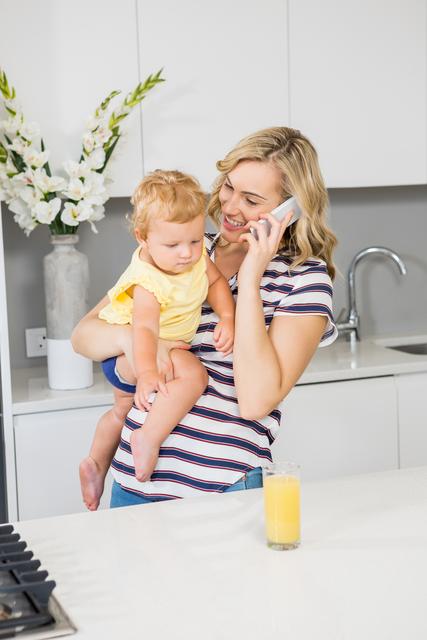 Mother holding baby girl while talking on mobile phone in kitchen. Ideal for themes related to parenting, multitasking, family life, modern motherhood, and home activities.