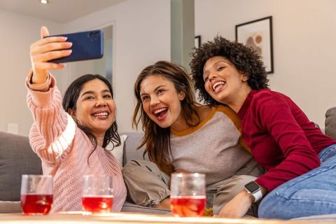 Multicultural women laughing and taking selfie on cozy living room sofa