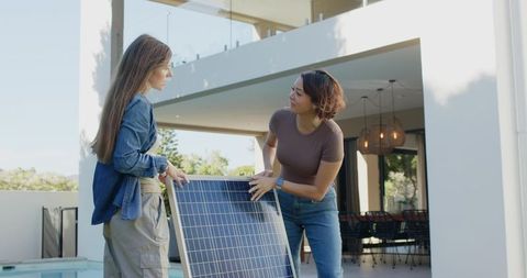 Two diverse women installing and inspecting solar panel poolside at modern home