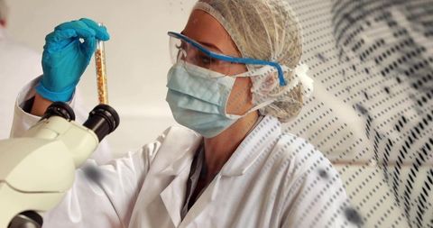 Female scientist inspecting vial under microscope wearing ppe mask gloves goggles