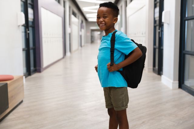 This image shows a cheerful African American elementary schoolboy standing in a school corridor with a backpack. Ideal for use in educational materials, back-to-school promotions, and advertisements focusing on childhood and learning.