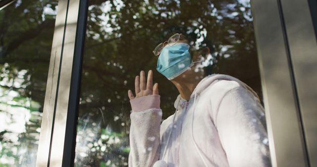 Young woman wearing a face mask looking through window while touching the glass. Reflections of trees visible in the background. Could be used for concepts involving pandemic, safety measures, isolation, mental health, and quarantine.