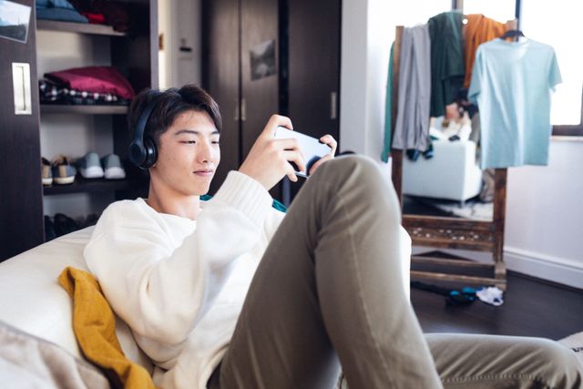Asian teenage boy sitting on couch, wearing headphones, playing game on smartphone. Background shows clothes and wardrobe, indicating a casual home environment. Ideal for use in articles about teenage lifestyle, home entertainment, technology use among youth, or digital gaming.