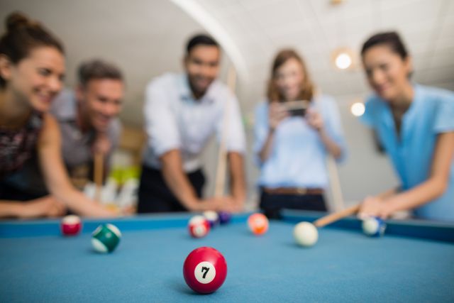 Business colleagues are gathered around a pool table, enjoying a game during break time in an office environment. This image can be used to illustrate teamwork, relaxation, and casual moments in a professional setting. Ideal for articles or advertisements focusing on workplace culture, team building activities, and employee engagement.