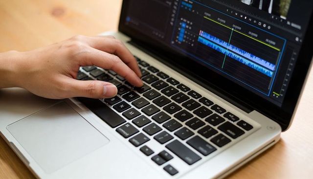 Hand editing video on metal laptop keyboard with visible audio waveform timeline