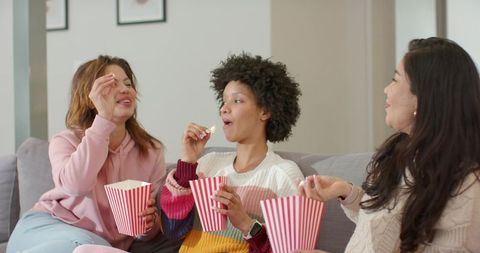 Three diverse women enjoying movie night on sofa wearing cozy sweaters and eating popcorn