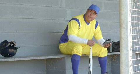 Baseball player in yellow uniform sitting in dugout holding bat and checking device