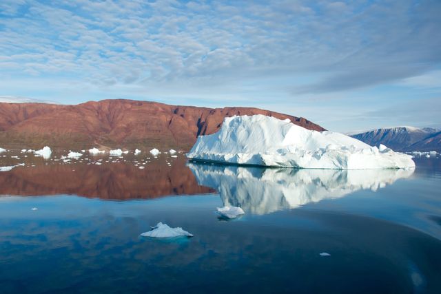 Iceberg floating on calm Arctic waters with a clear reflection, red mountains in the background under a partly cloudy sky. Ideal for use in environmental awareness campaigns, travel brochures, nature documentaries, and educational materials focusing on climate change and polar regions.