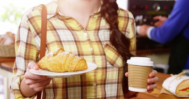 Young woman in plaid shirt holding coffee in to-go cup and croissant on plate, standing in café. Great for depicting casual morning routine, enjoying breakfast, or promoting café and bakery products.