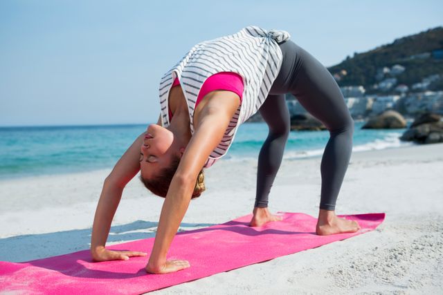 Young woman practicing yoga in bridge position on a beach during a sunny day. Ideal for promoting fitness, wellness, and healthy lifestyle content. Can be used in articles, blogs, or advertisements related to outdoor activities, yoga retreats, and relaxation techniques.