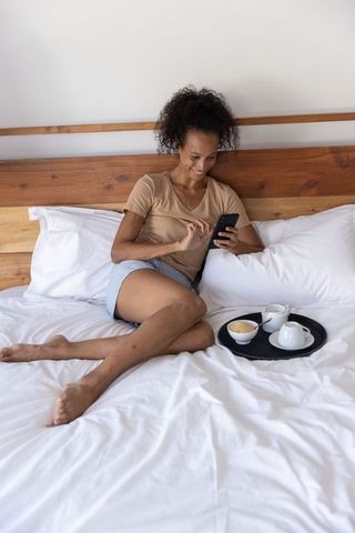 Relaxing woman on bed checking smartphone with breakfast tray and cozy linens