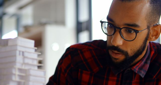 Young architect wearing glasses and plaid shirt deeply focused on a scale model in a modern office. Ideal for showcasing architecture professions, creative workspaces, office culture, and professional dedication.