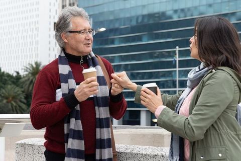 Senior man and asian woman chatting over takeaway coffee on city terrace by office tower