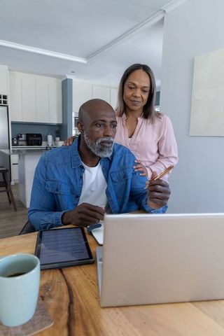 Senior couple working together on laptop and tablet at dining table in modern kitchen