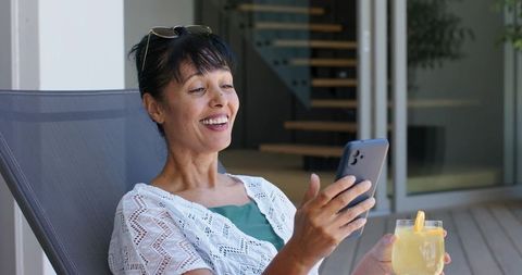 Middle-aged woman relaxing on modern terrace, smiling at smartphone while holding lemonade glass
