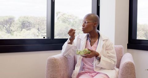 African Woman Doctor Eating Salad During Clinic Break, Sitting Relaxing in Sunlit Lounge