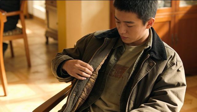Teen male adjusting brown zip-front jacket with corduroy collar, sitting in sunlit room