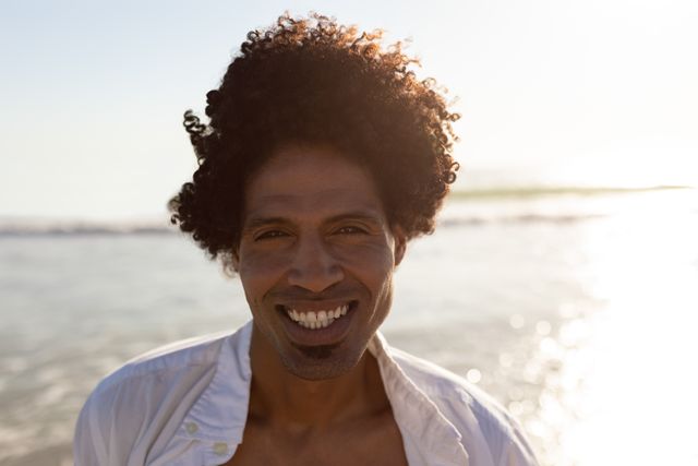 Young man with curly hair smiling while standing on a sunny beach. Ideal for use in advertisements, travel brochures, lifestyle blogs, and social media posts promoting outdoor activities, vacations, and positive emotions.