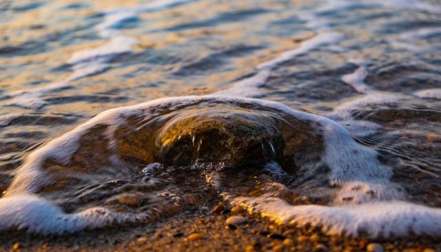 Golden-hour waves washing rounded shoreline rock with foamy surf and glistening pebbles