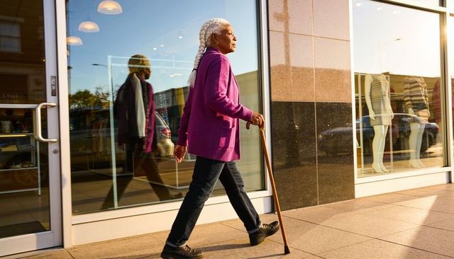 Senior woman walking with wooden cane past retail windows wearing magenta cardigan