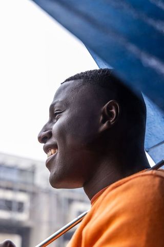 Smiling african american man holding blue umbrella and wearing orange shirt in rainy city