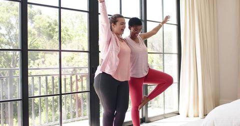 Two women practicing partner yoga balancing in bright bedroom with large black-framed doors
