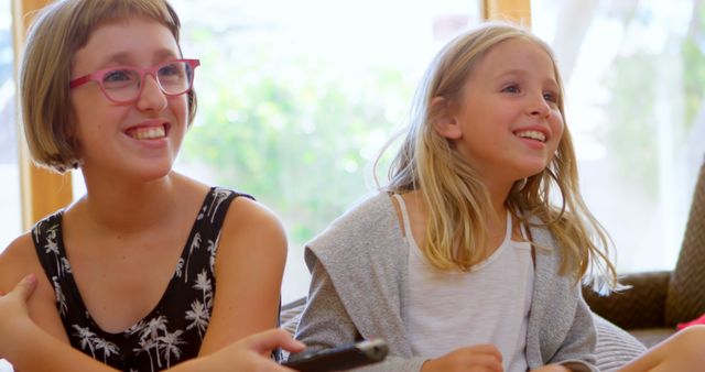 Teenage Caucasian girl and Caucasian girl enjoy a bright home setting. They share a joyful moment, watching something entertaining.