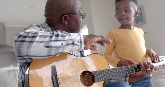 Grandfather teaching his young grandson how to play an acoustic guitar in a cozy living room setting. The image captures a heartwarming moment of familial bonding and the transfer of knowledge between generations. Suitable for use in family-oriented advertisements, educational materials about music, or articles about intergenerational relationships and learning.