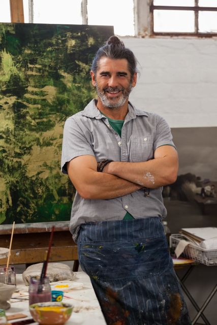 Middle-aged male artist standing confidently with arms crossed in a creative studio. He is wearing a casual shirt and an apron, surrounded by art supplies and paintings. Ideal for use in articles about creativity, artistic professions, and personal inspiration.